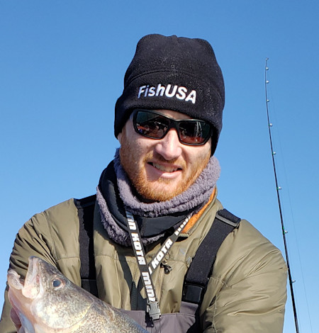 Fisherman in black beanie labeled FishUSA, sunglasses, holding a fish with rod behind