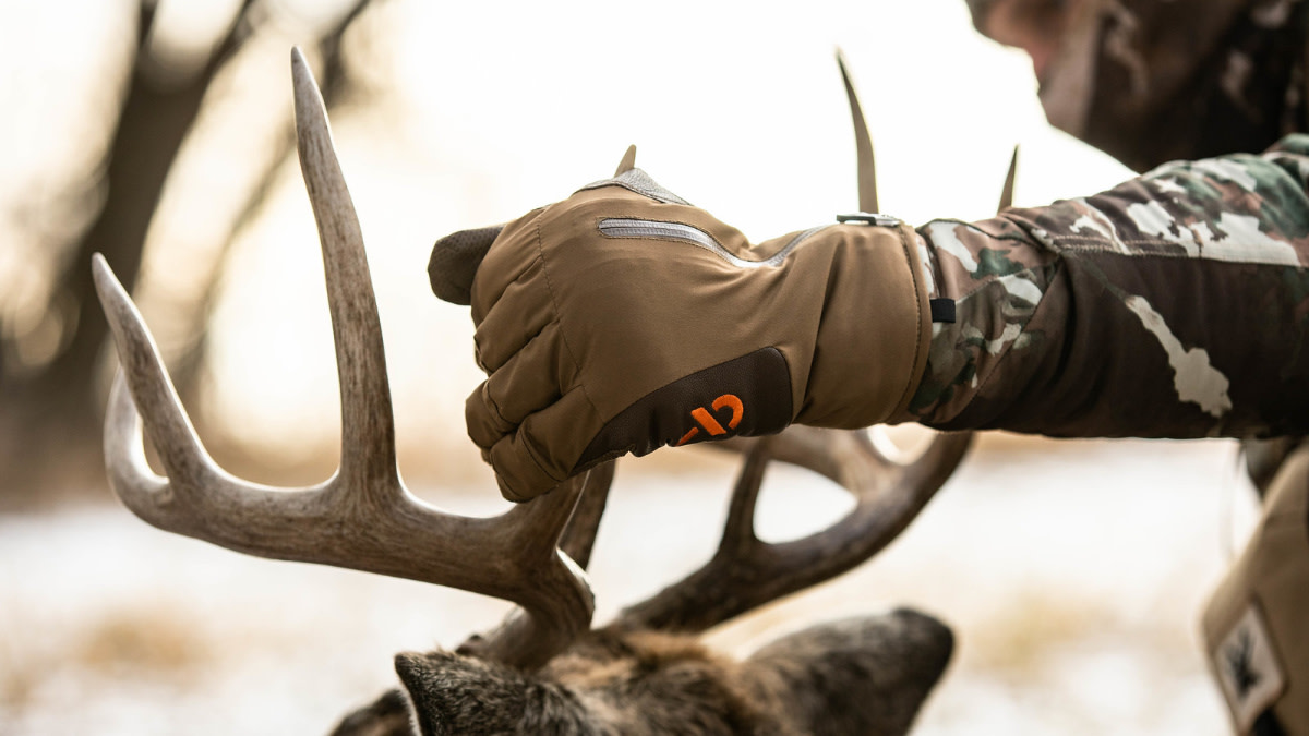 Gloved hand holding buck antlers above deer head, camo sleeve and snowy blurred background