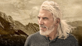 Older man with long white hair and beard, facing right; mountains and faded map behind him