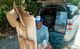 Artist painting at easel beside open SUV, straw hat with feathers on easel, painted canvases in trunk.