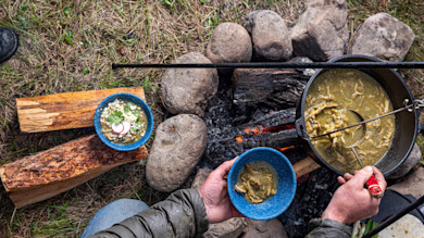 Person ladling green chili stew from a Dutch oven into a blue enamel bowl over a campfire