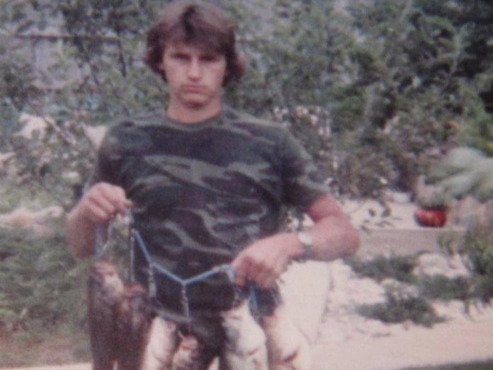 Young man in camo shirt holding a blue stringer of fish with shrubs behind