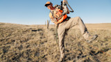 Hunter in orange vest and cap stepping over a barbed-wire fence carrying a rifle on prairie