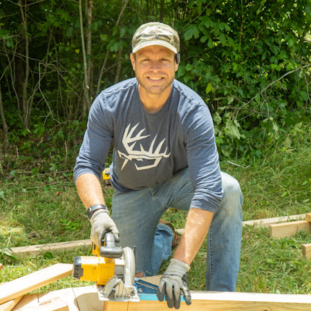 Man kneeling cutting lumber with a circular saw, wearing blue shirt with white antler logo