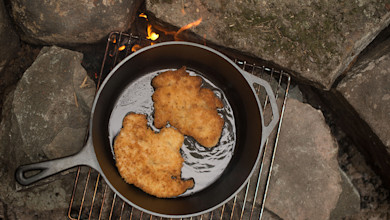 Cast-iron skillet frying two breaded turkey schnitzels over a campfire on a grill grate