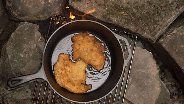 Cast-iron skillet frying two breaded turkey schnitzels over a campfire on a grill grate