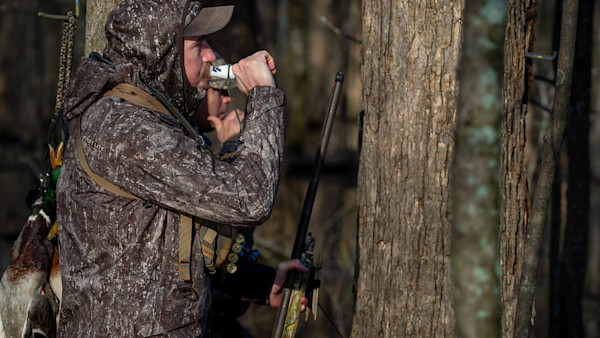 Hunter in First Lite camo jacket blowing white duck call beside shotgun and hanging mallards