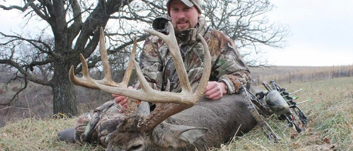 Hunter kneeling behind large buck with massive antlers, compound bow and arrows on grass