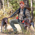 Hunter wearing orange cap kneeling in woods with shotgun over shoulder and two hunting dogs