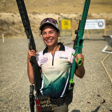 Woman shooter at outdoor range, smiling, holding two green rifles, wearing cap and shirt labeled "TAYLOR"