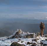 Hunter with backpack and rifle standing on snowy ridge overlooking mountain valley