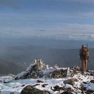 Hunter with backpack and rifle standing on snowy ridge overlooking mountain valley