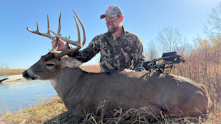 Hunter kneeling behind large buck at riverbank holding antlers; compound bow rests on deer