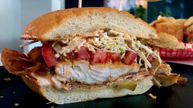 Fried walleye po'boy sandwich with coleslaw, tomato, pickles, bacon and spicy mayo