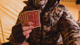 Hunter in camouflage jacket and fingerless glove holding five red-backed playing cards inside a tent
