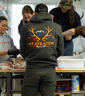 Person seen from back wearing hoodie with orange MEATEATER 'Bozeman, MT' logo; people cutting meat