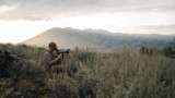 Hunter kneeling in sagebrush using a spotting scope on tripod with mountains at sunrise