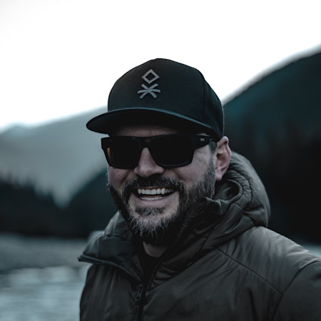 Smiling man in sunglasses, black cap and puffer jacket standing by a river with mountains