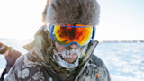 Man with ice-crusted mustache wearing fur hat, mirrored goggles and camouflage jacket in snow