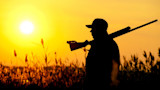 Hunter silhouetted at sunset carrying shotgun over shoulder in tall grass
