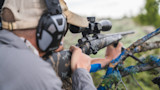Hunter aiming scoped rifle resting on a tree branch, wearing ear protection and cap