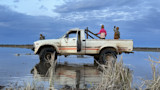 Old white pickup truck in shallow water with open door; man at front and two children standing in truck bed