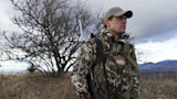 Hunter in camouflage jacket and cap with rifle slung, standing by bare tree and distant mountains
