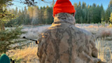Hunter seen from behind in orange beanie and camo jacket, rifle pointed into frosty meadow