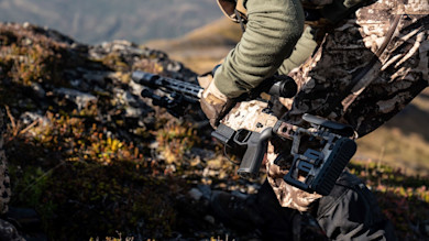 Hunter kneeling on tundra holding camouflaged rifle with scope and bipod