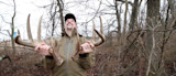 Man holding a pair of large deer antlers, laughing in leafless woods
