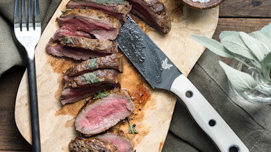 Medium-rare sliced venison steak with sage on wooden board, chef's knife and fork