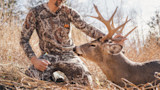 Hunter kneeling in First Lite camouflage, holding a harvested whitetail buck by its antlers