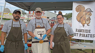 Three people in aprons under a tent with banner "RAWHIDE NUT PIRATES", center man holds small game