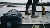 Person standing in aluminum boat wearing muddy rubber boots, with paddle, dry bag and coiled rope