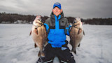 Man holding two large fish on frozen lake, kneeling on ice with treeline background