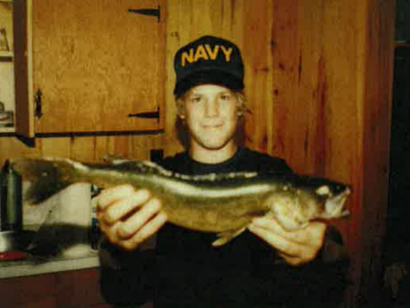 Man holding fish in kitchen, wearing black cap reading 'NAVY'