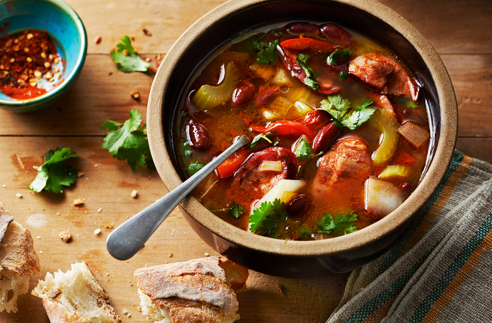 Un ragoût à l'espagnole au chorizo et aux haricots rouges servi dans un petit bol à soupe en céramique brune. Le ragoût est garni de persil haché et accompagné de morceaux de pain grillé pour tremper