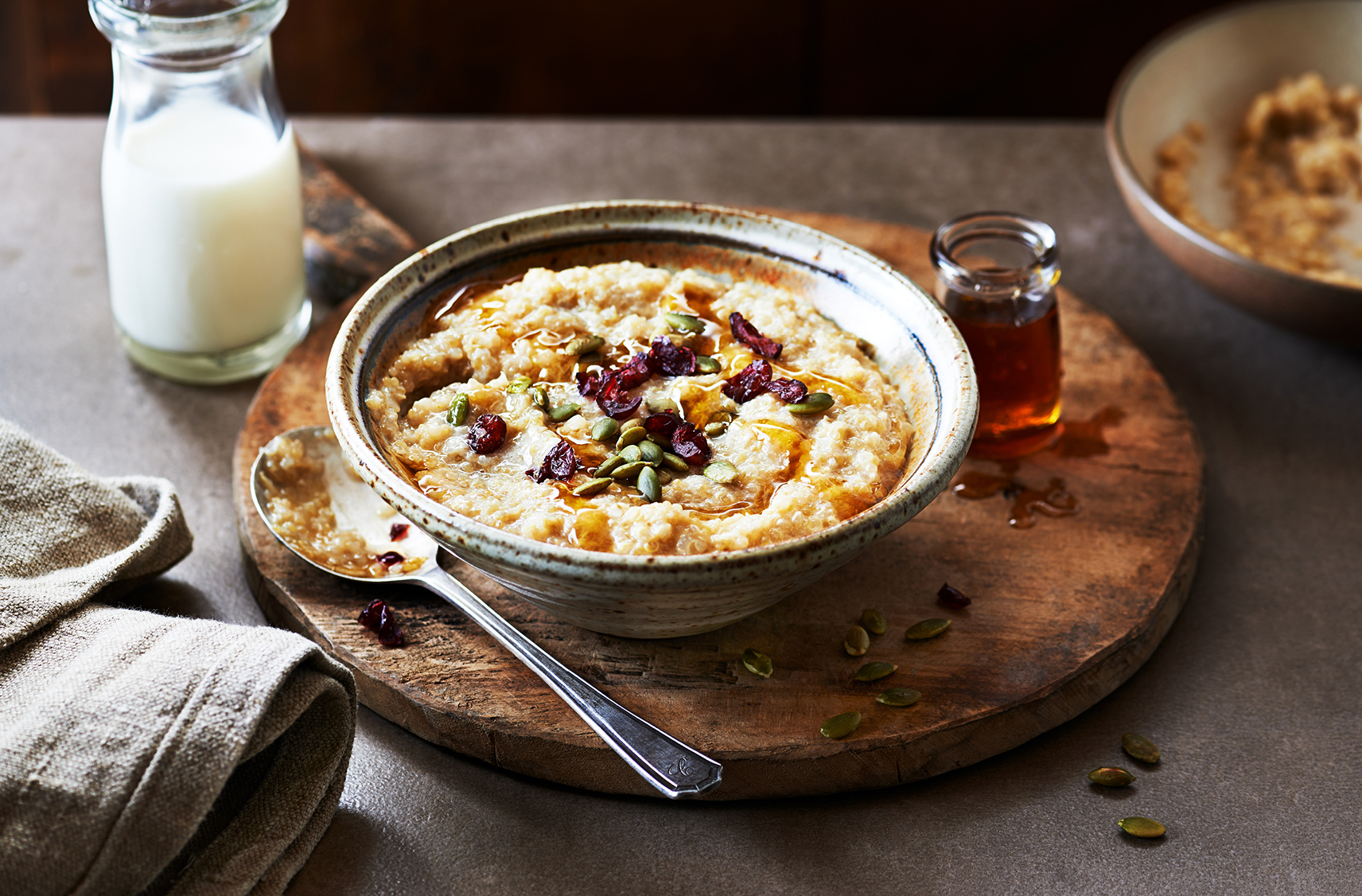 Bowl of porridge with pumpkin seeds and cranberries on top on a wooden round cutting board next to a spoon with a glass of milk
