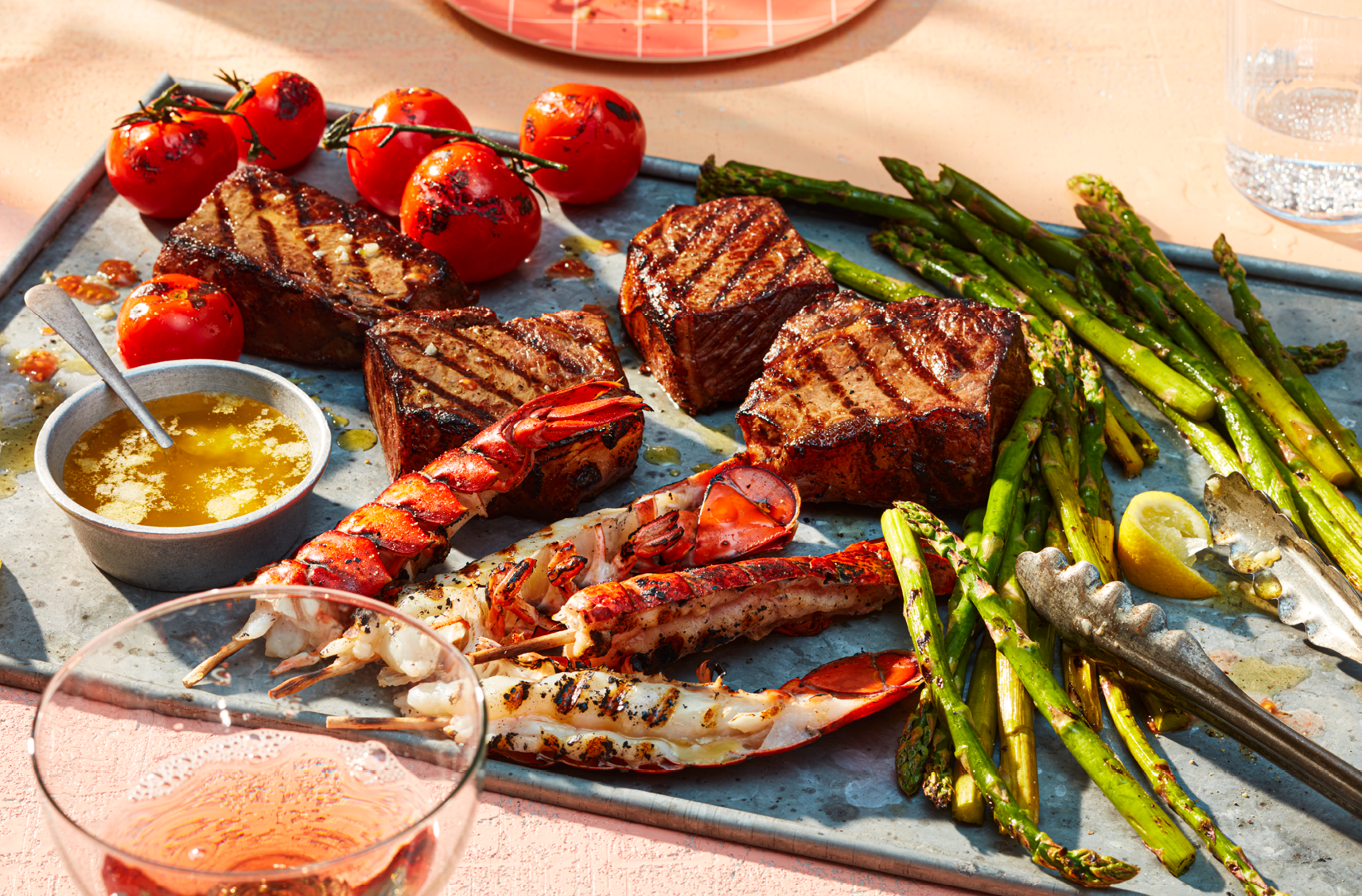 A tray filled with grilled portions of PC Lobster Tail Skewers, striploin steak, blistered tomatoes and asparagus tips