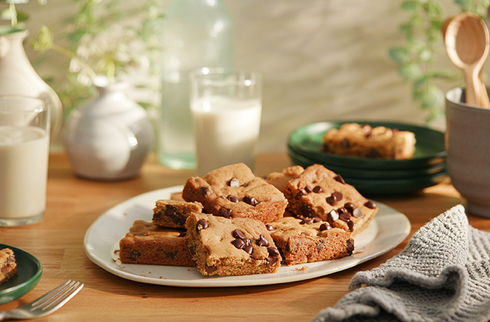A plate filled with plant based vegan chocolate chip blondies on a kitchen table with glasses of milk beverages and utensils