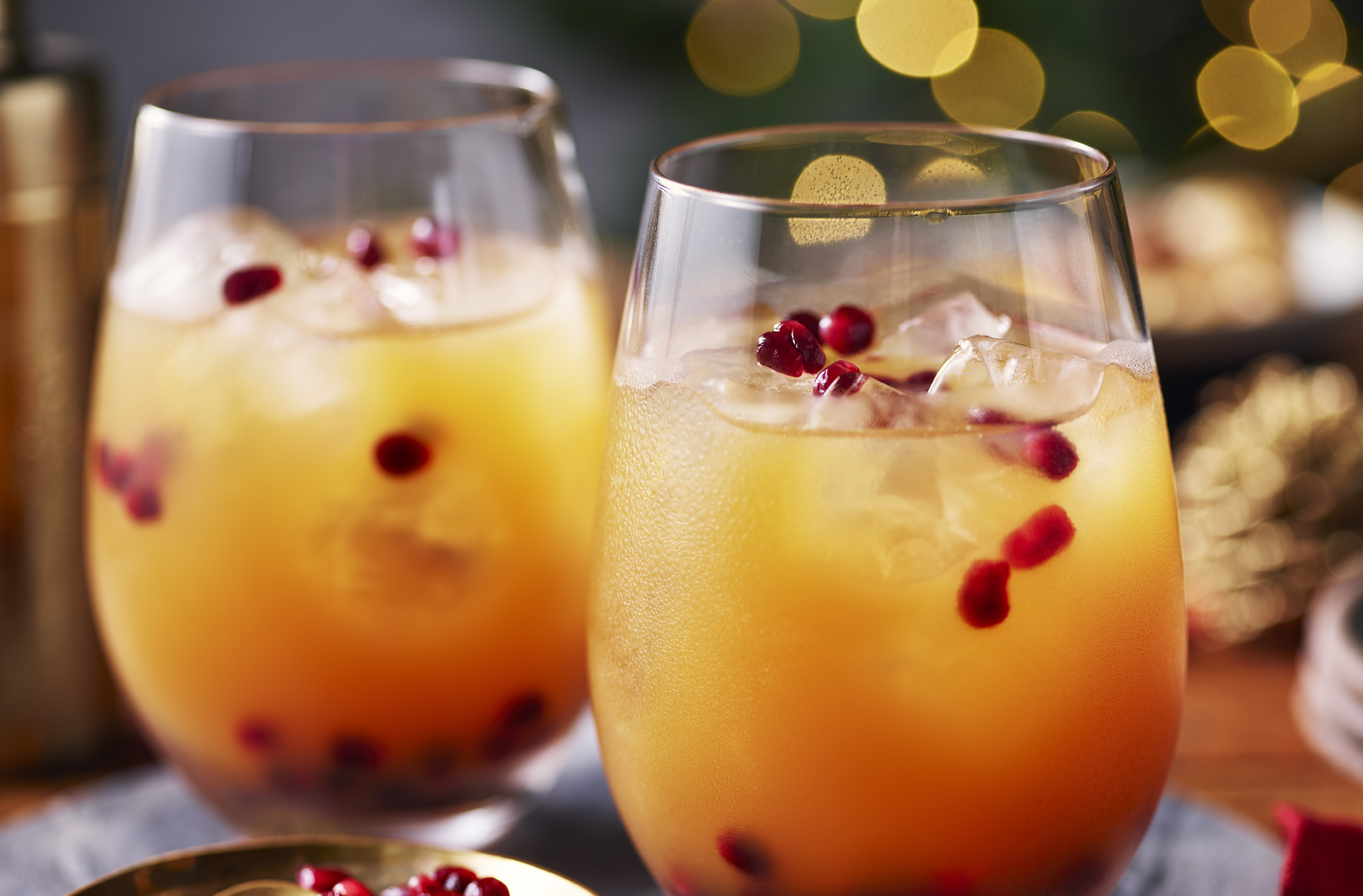 Two golden juices with ice and pomegranate seeds rest beside more seeds
