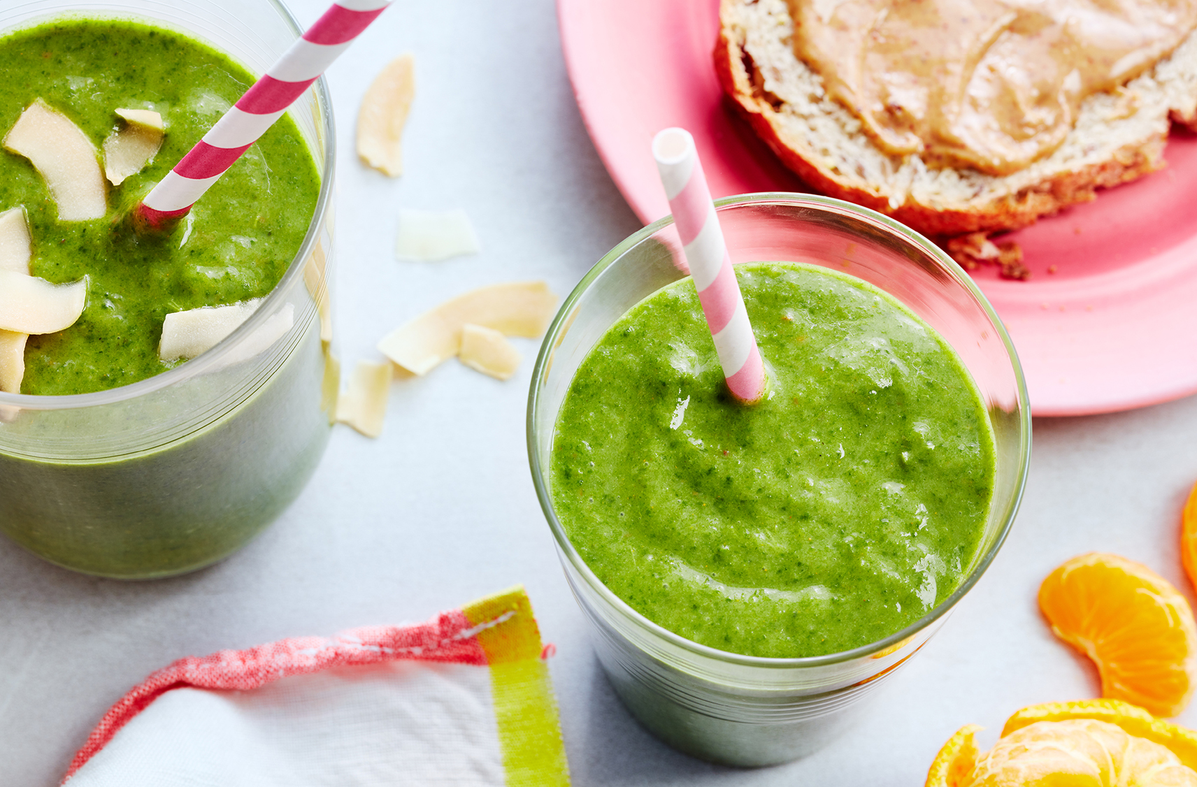 A slice of bread with nut butter sits beside two green smoothies with straws