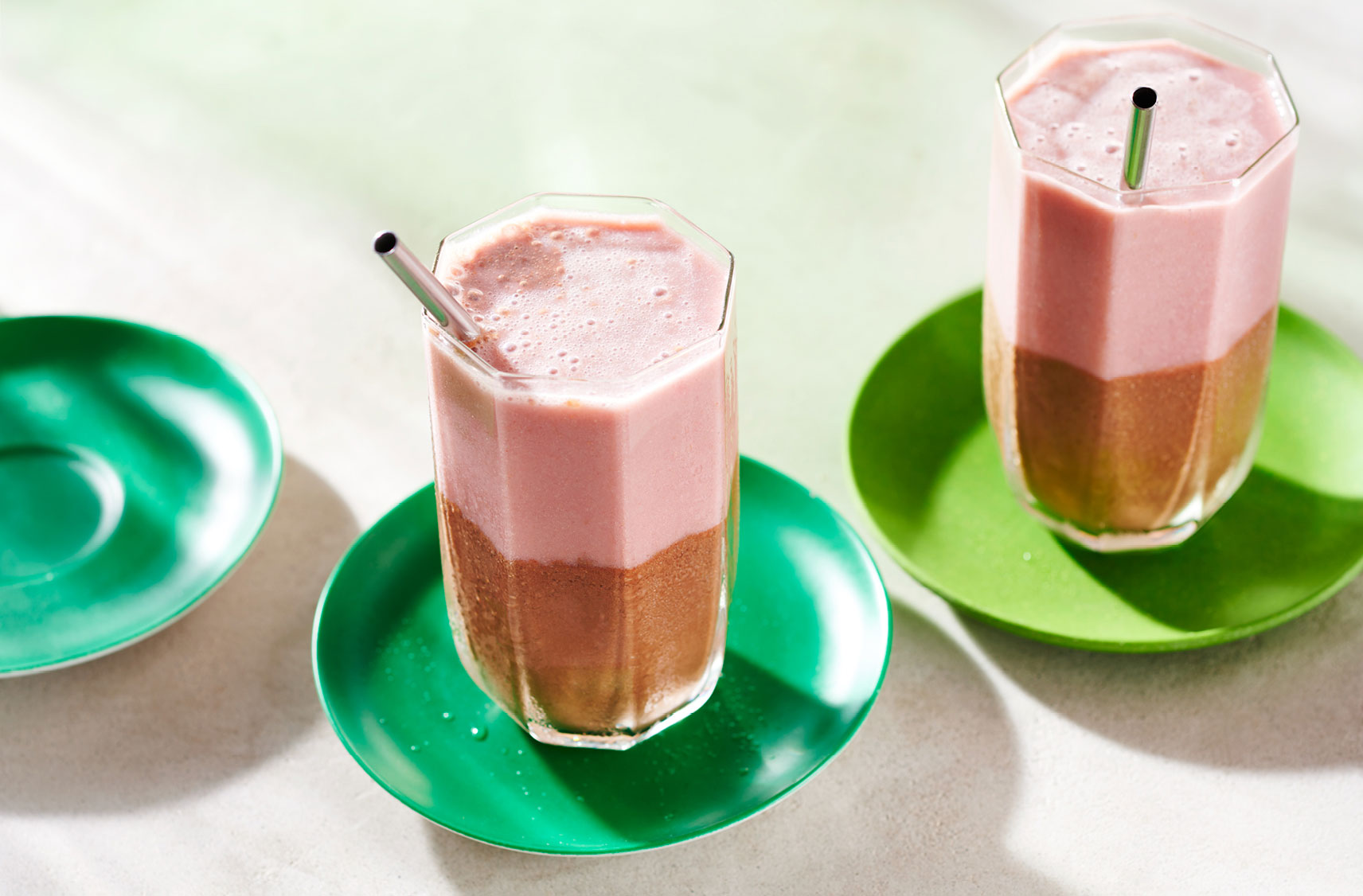 Trois petites soucoupes, dont deux contiennent des smoothies étagés aux fraises et au chocolat avec des pailles réutilisables.
