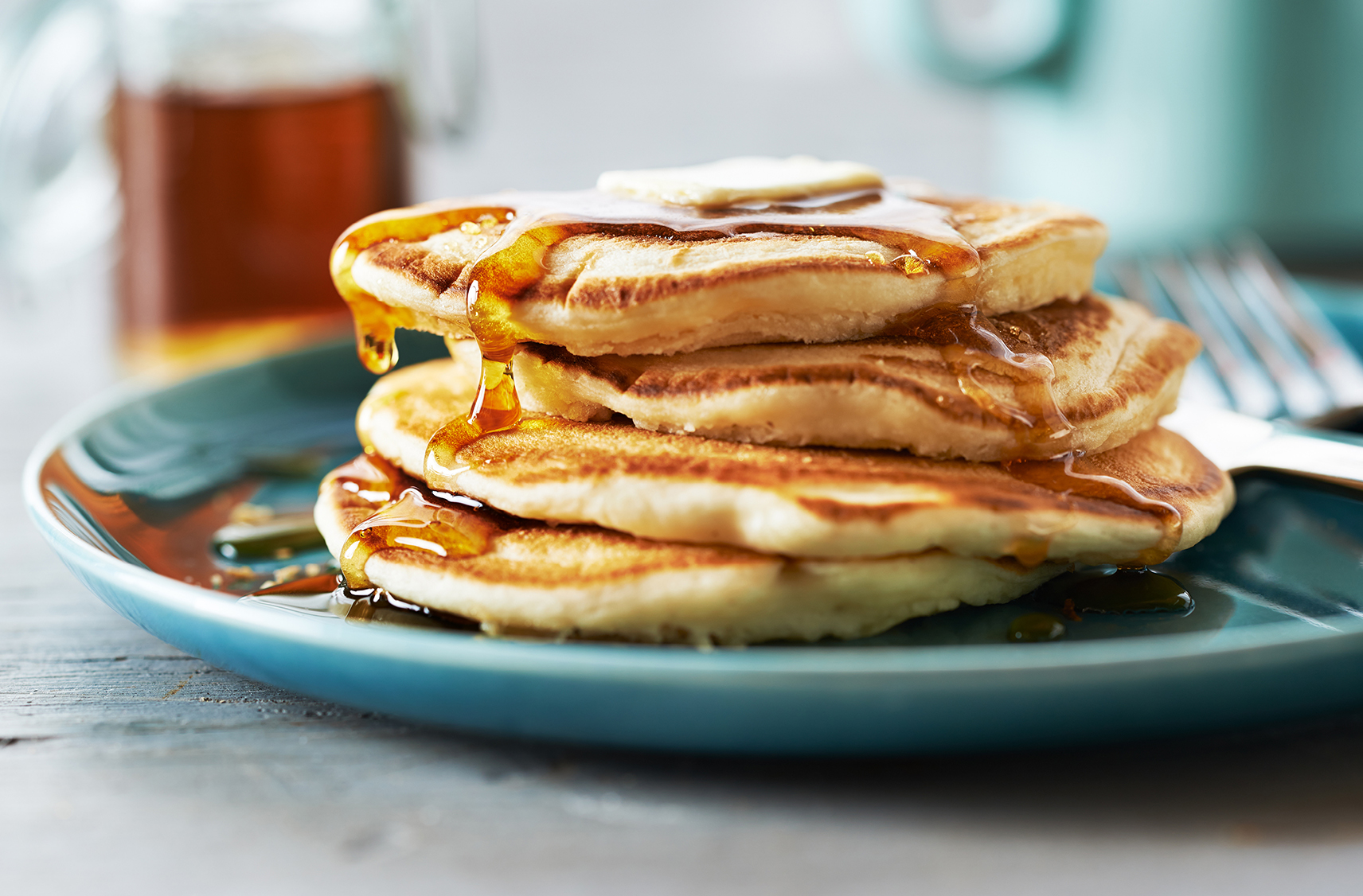Stack of 4 buttermilk pancakes on a plate, topped with butter & maple syrup