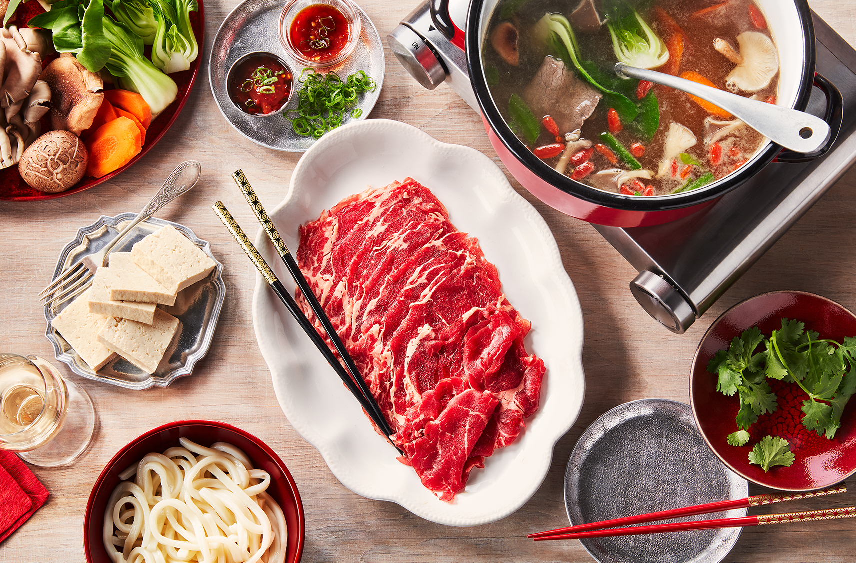 Le dessus d'une table avec plusieurs plats pour un hot pot chinois, dont du bœuf au centre et plusieurs assiettes de légumes et autres à-côtés.