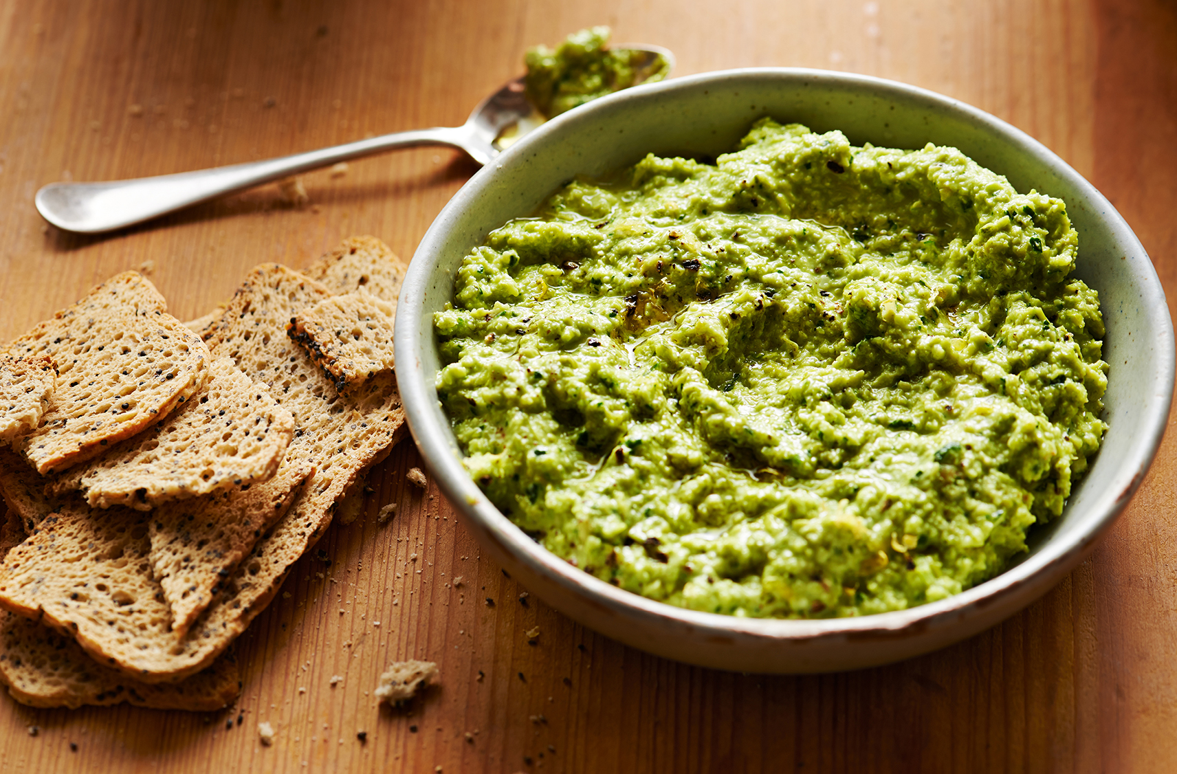 green hummus served in a small grey bowl with slices of dry toast on the side for dipping