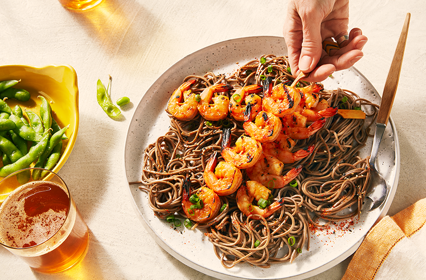 A dish of Spicy soba noodles with 3 grilled orange miso shrimp skewers being placed on top.