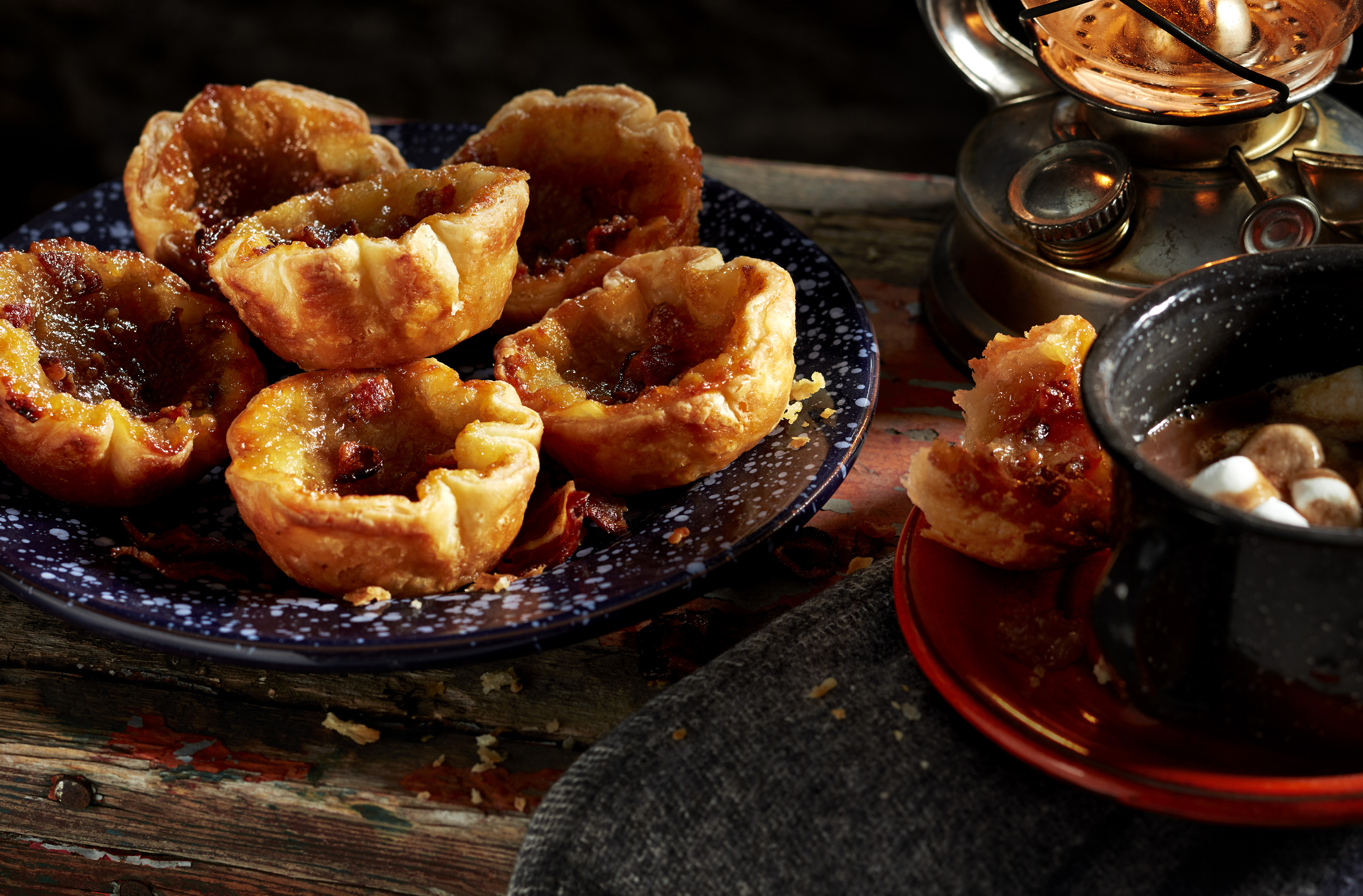 Six butter tarts stacked on a plate beside a lantern and hot chocolate