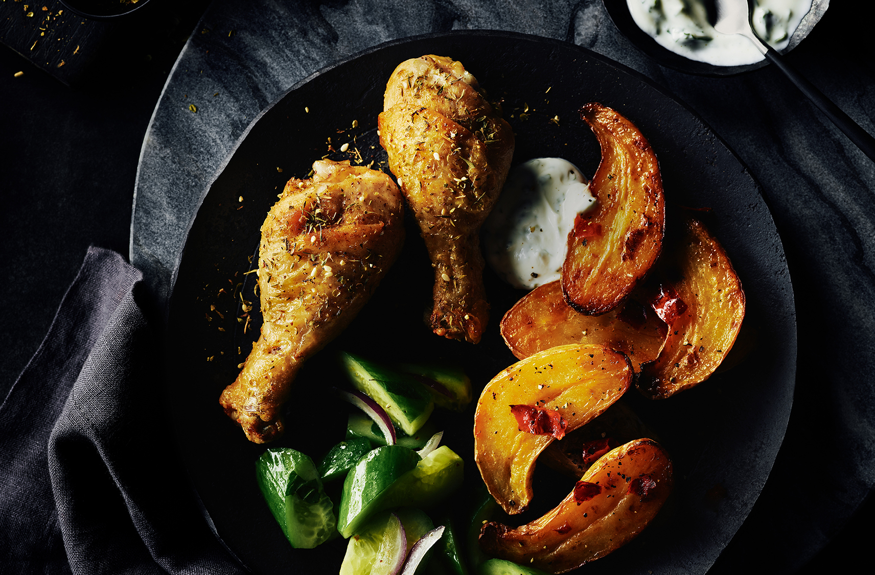 two pieces of zaatar chicken drumsticks on a black dinner plate served alongside spicy fingerling potatoes, cucumber chunks and a dollop of mint yogurt