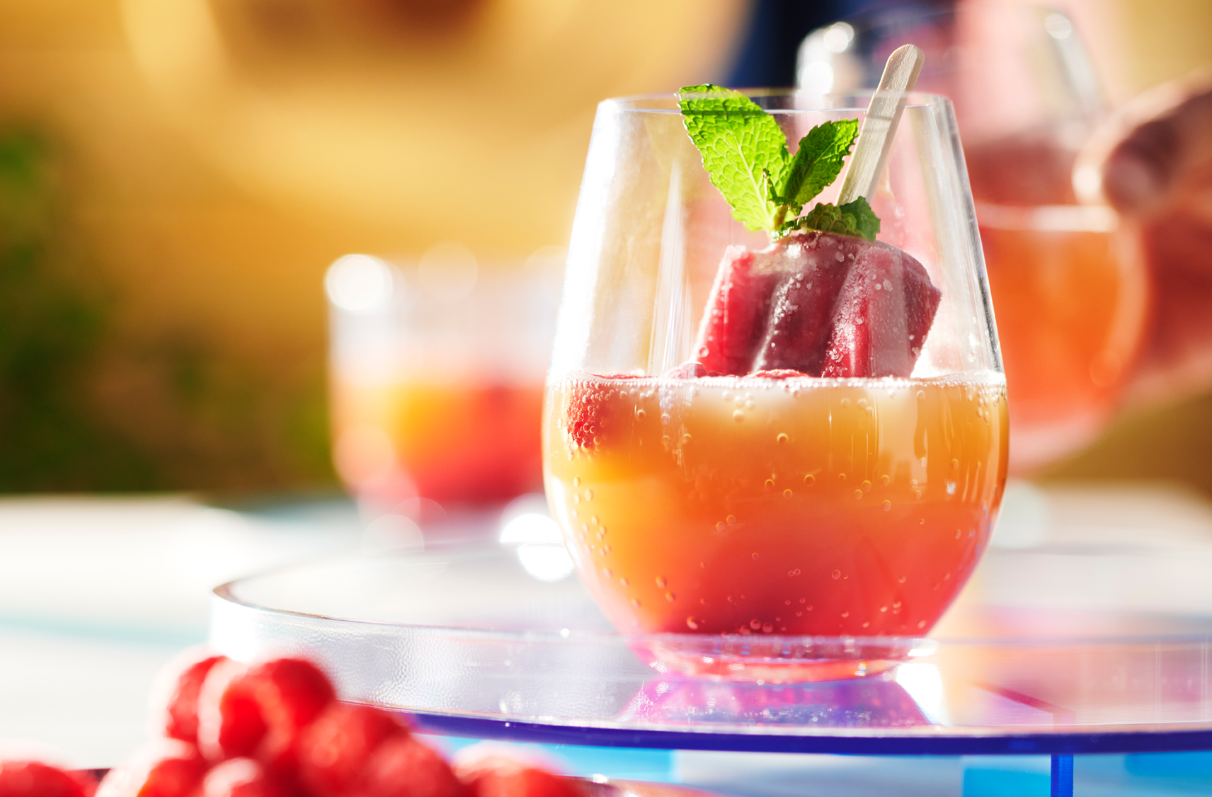 A hand dipping a berry coloured ice pop into a glass filled with orange juice and a sprig of mint.